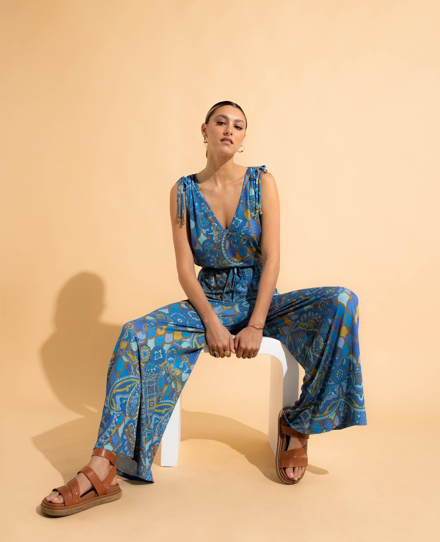 Woman wearing a blue patterned jumpsuit sitting on a white stool against a beige background