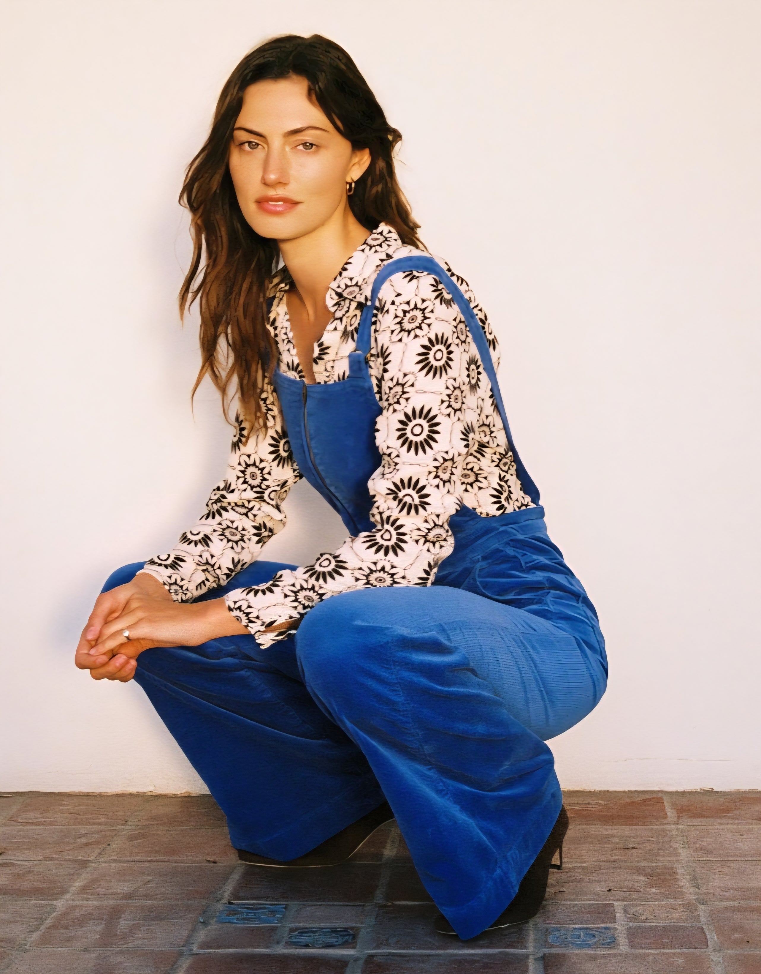 Woman wearing a floral blouse and blue overalls sitting on the floor against a white wall.