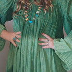 Woman wearing a green dress with puffed sleeves and a beige hat, posing with hands on hips.