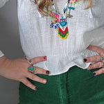 Person wearing a colorful necklace, white blouse, and green pants with a gray background.