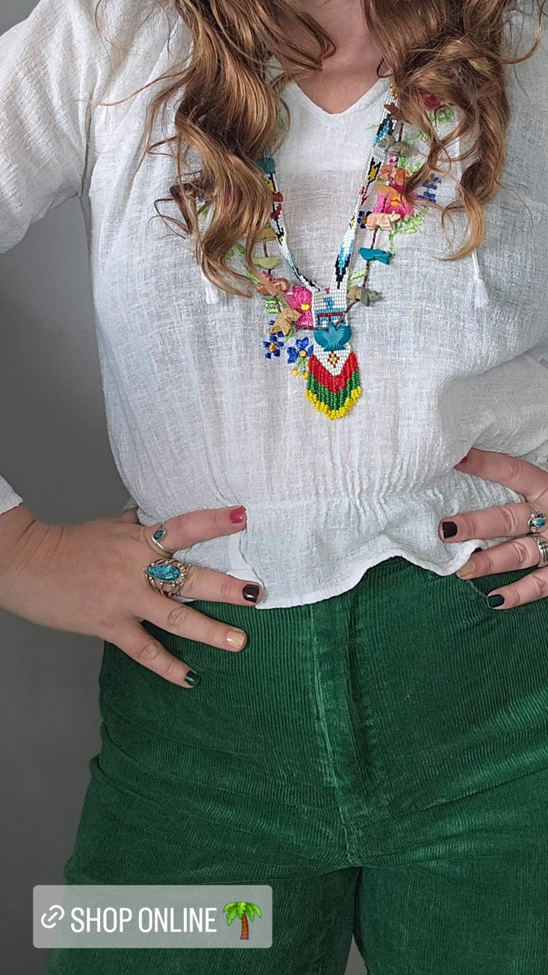 Person wearing a colorful necklace, white blouse, and green pants with a gray background.