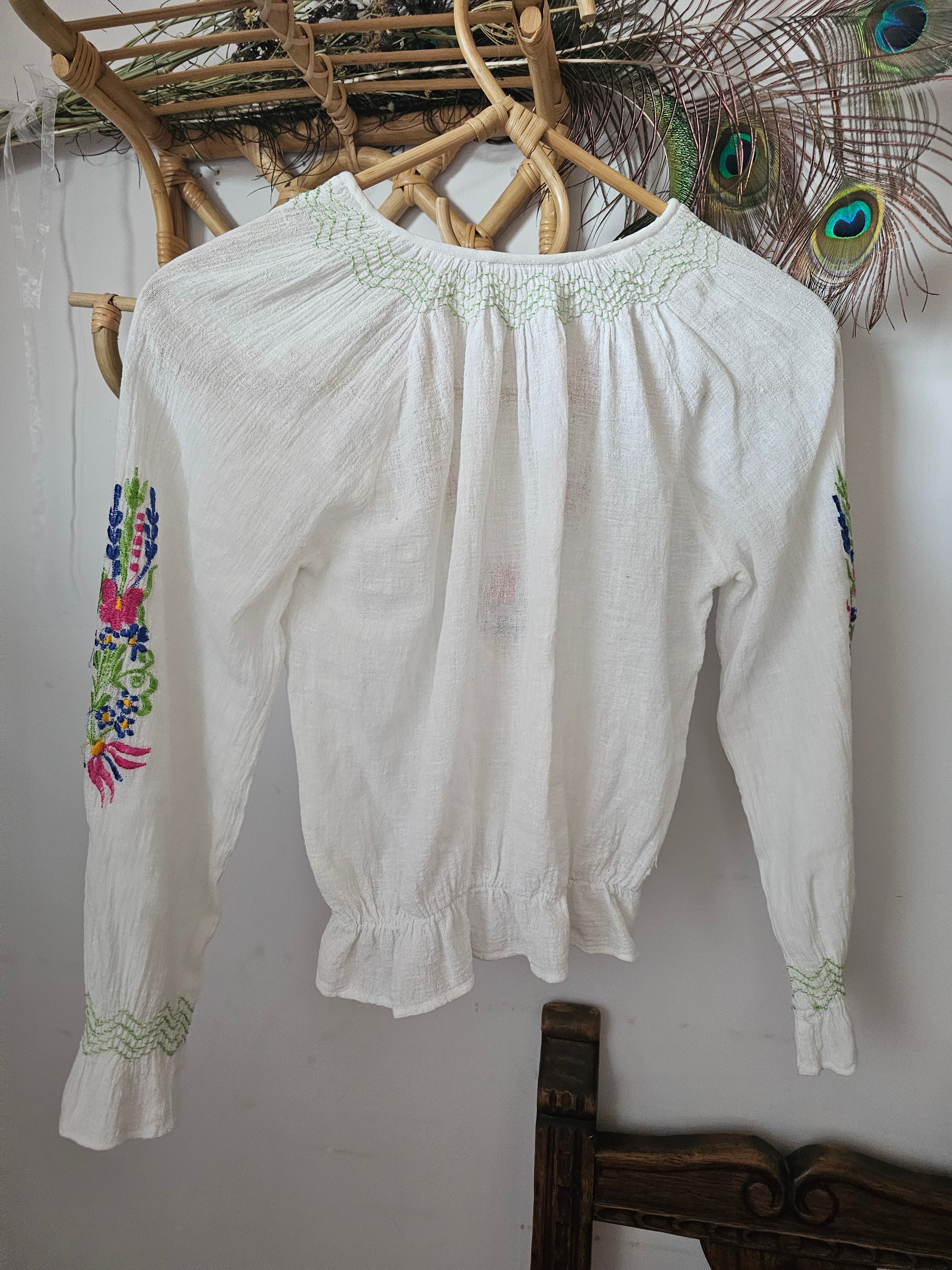 White blouse with floral embroidery on a white surface with a peacock feather decoration.