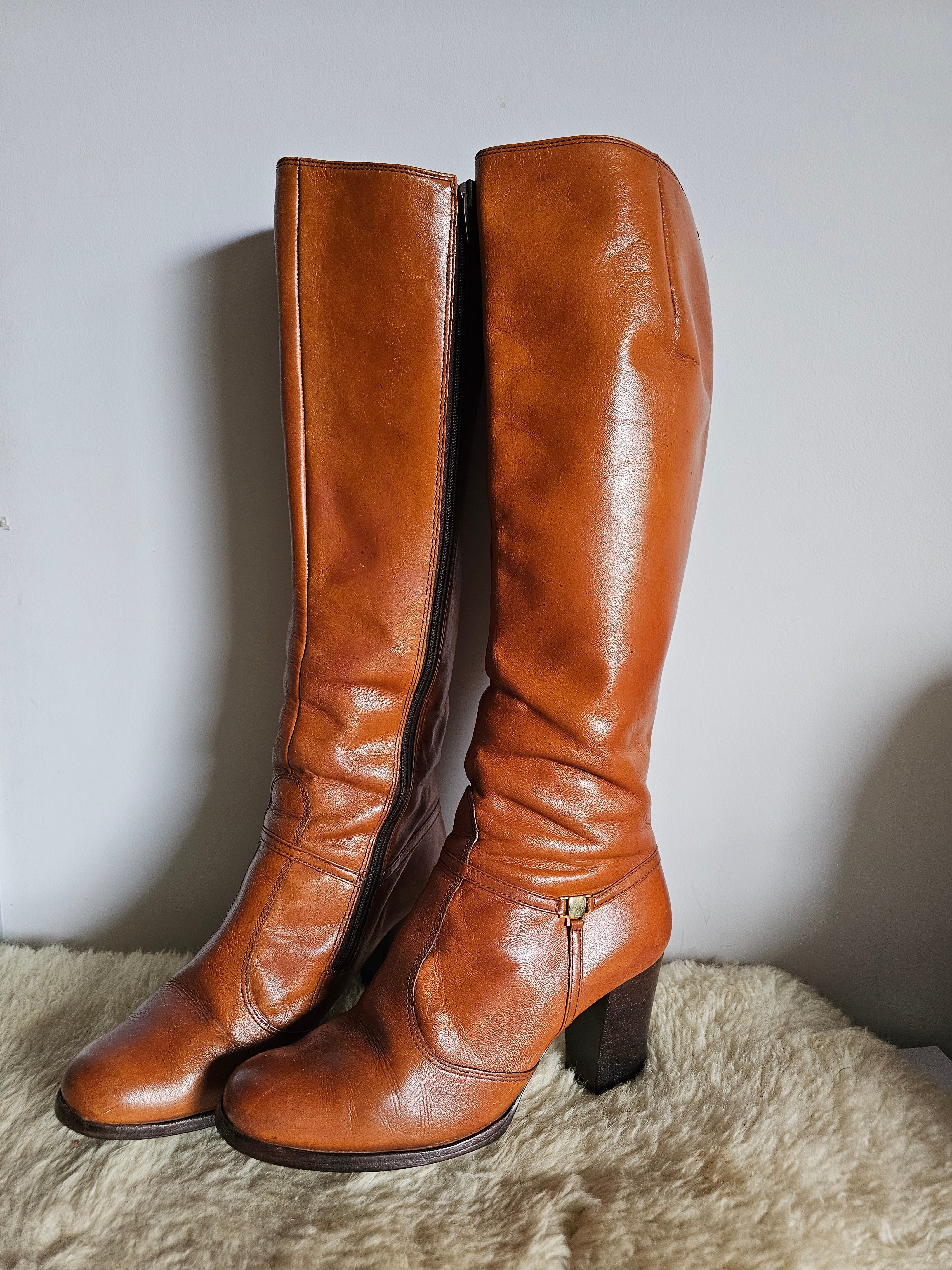 Pair of brown leather knee-high boots on a light gray surface with a textured beige fabric background.