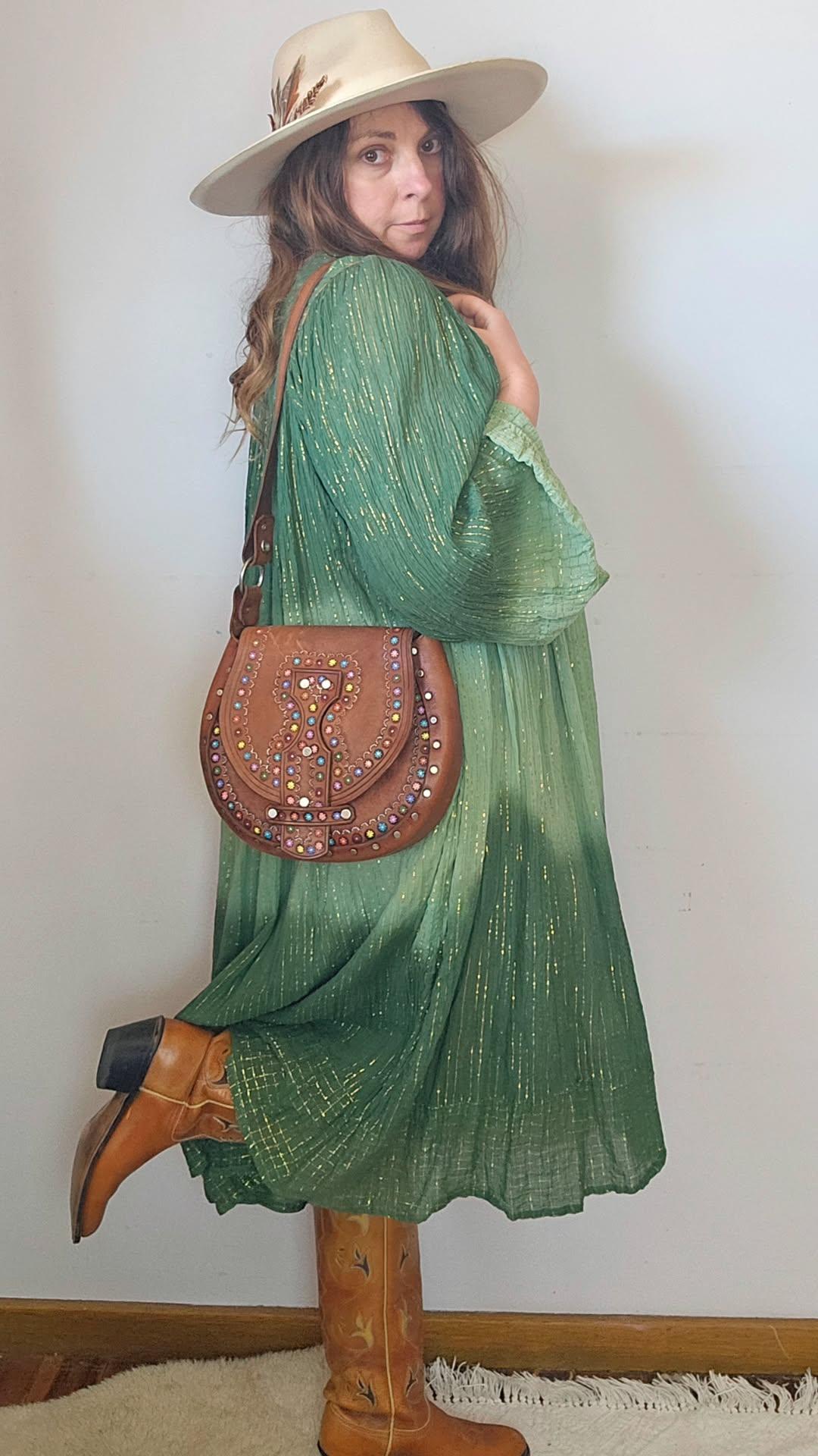 Woman in a green dress and brown boots holding a brown handbag against a plain background