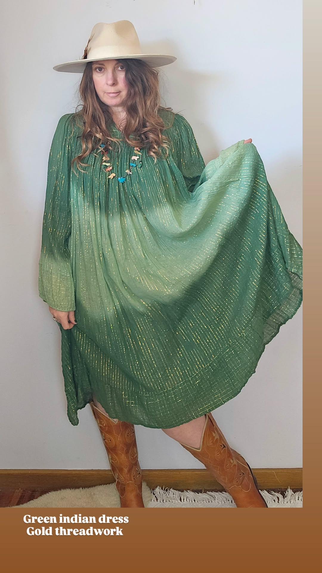Woman wearing a green dress with gold threadwork, standing against a white wall.