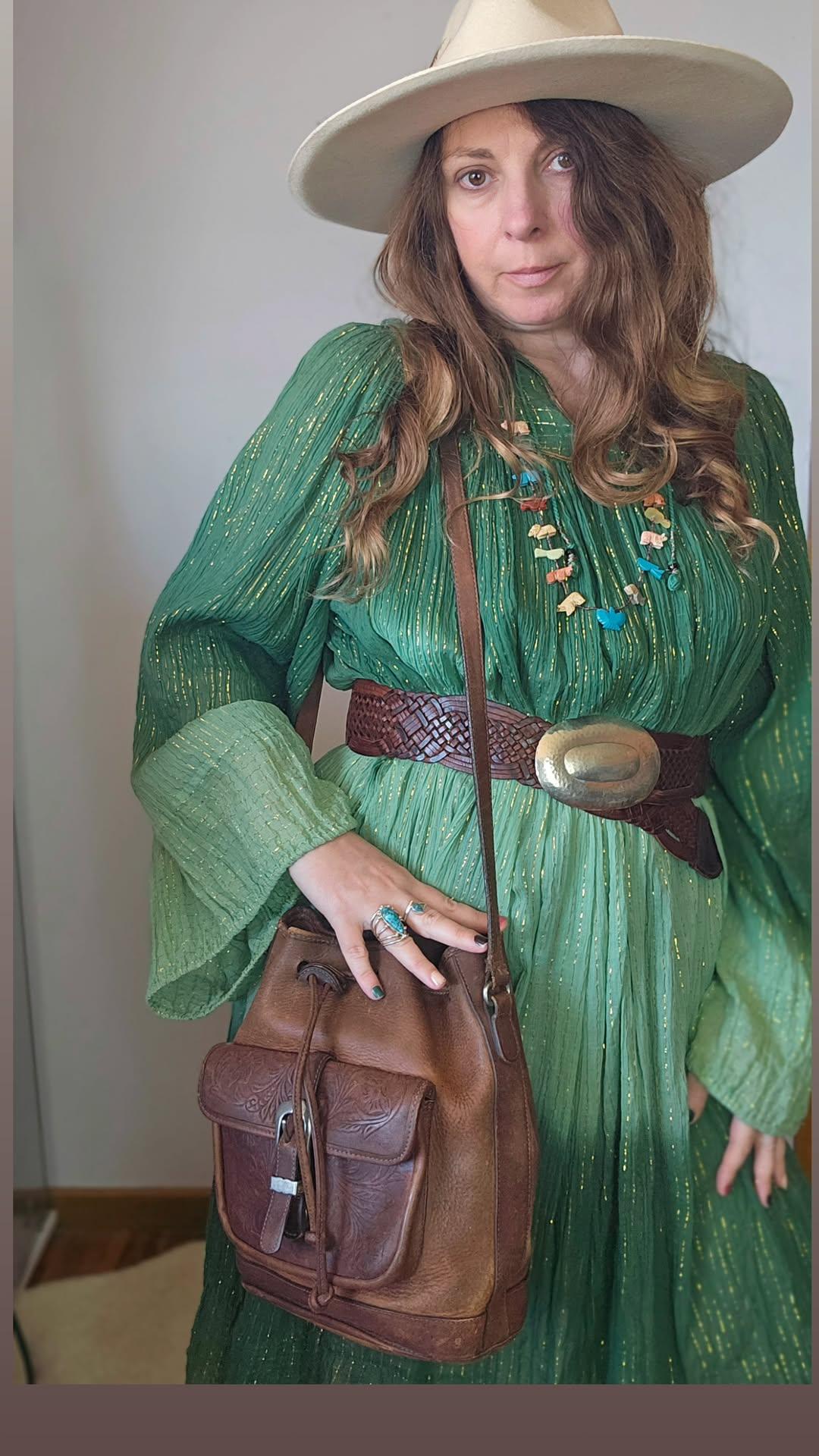 Woman in a green dress with a brown belt and a brown leather bag, wearing a beige hat.