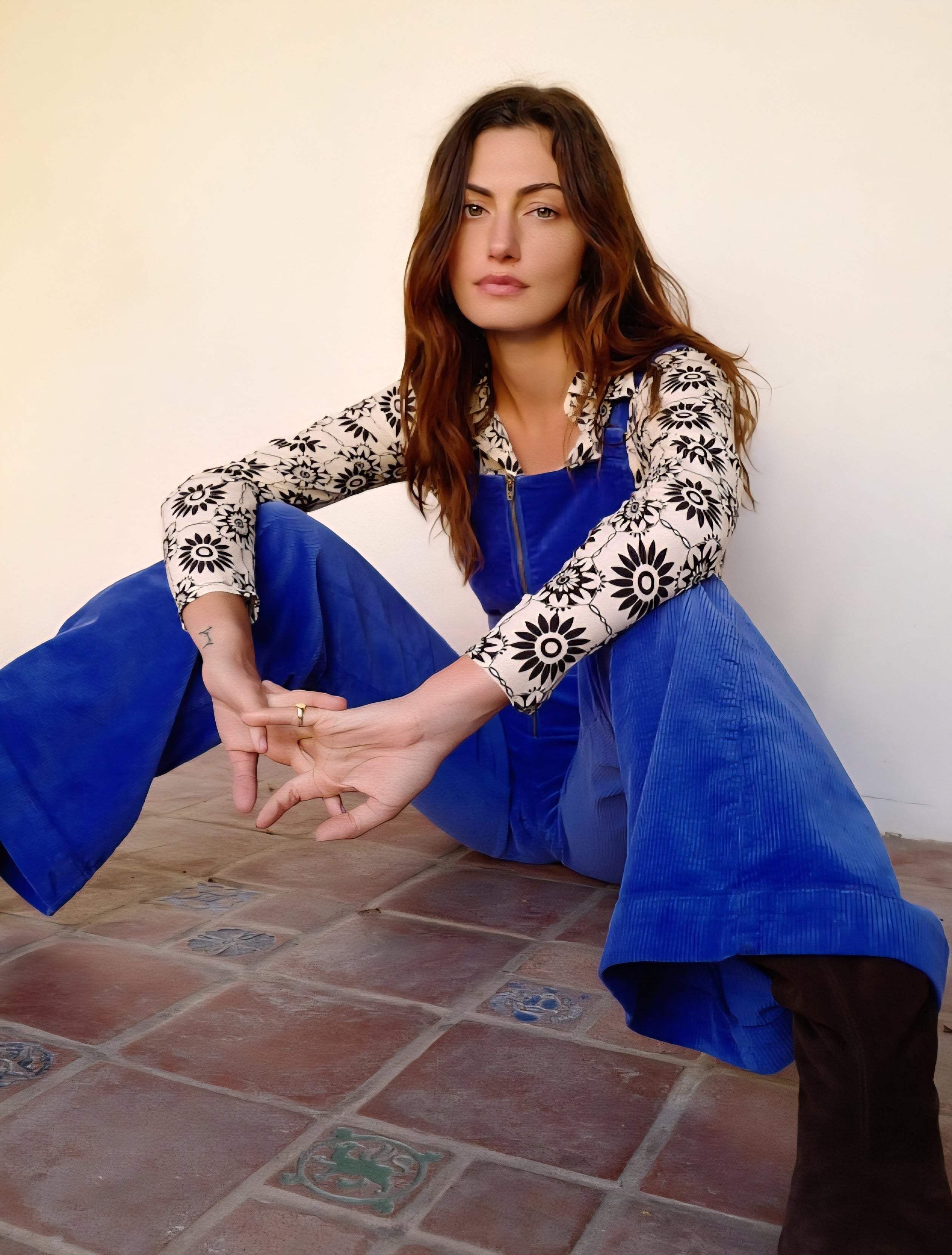 Woman sitting on a tiled floor wearing a blue dress and patterned top.