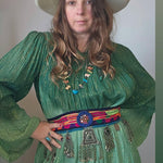 Woman wearing a green dress with a colorful belt and a white hat against a plain background.