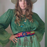 Woman wearing a green dress with a colorful belt and a white hat against a plain background.