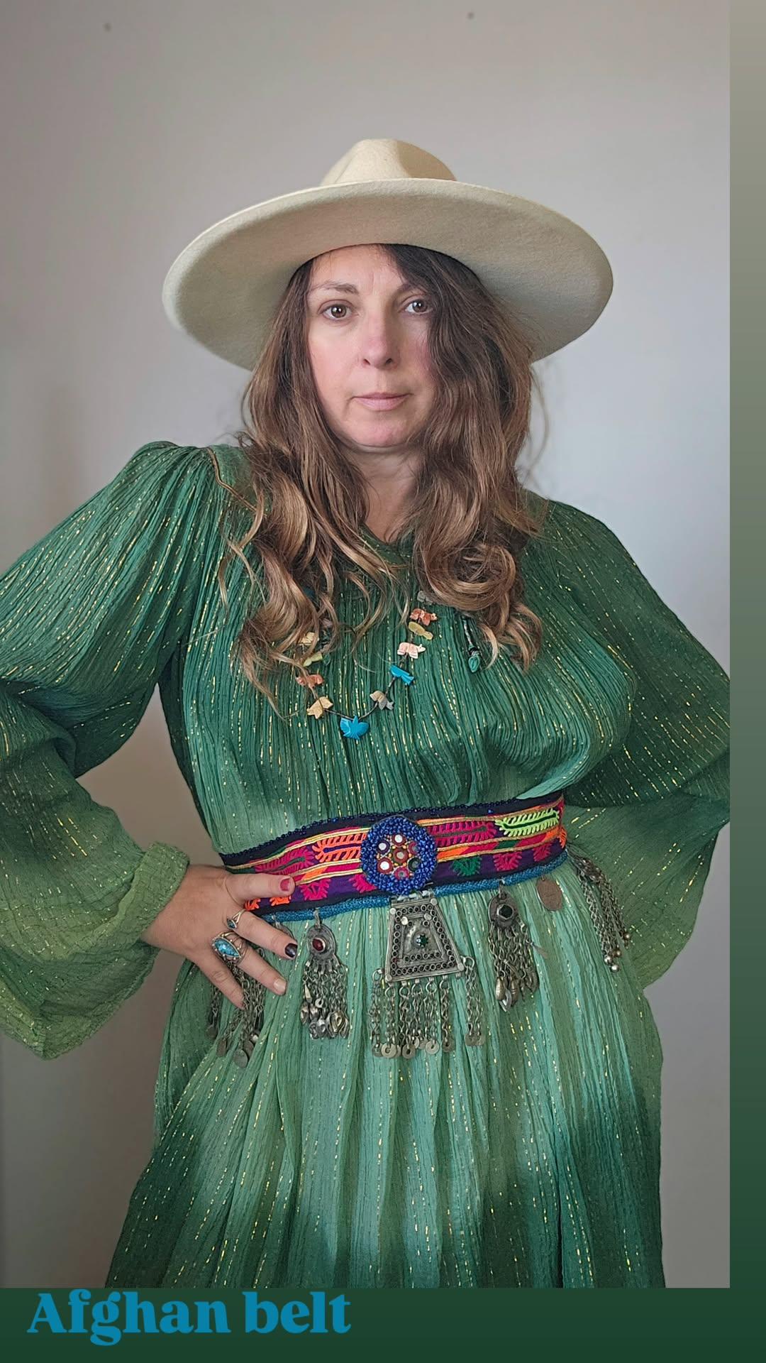 Woman wearing a green dress with a colorful belt and a white hat against a plain background.