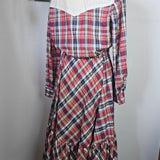 Plaid dress on a mannequin against a white wall with a framed picture and hat in the background.