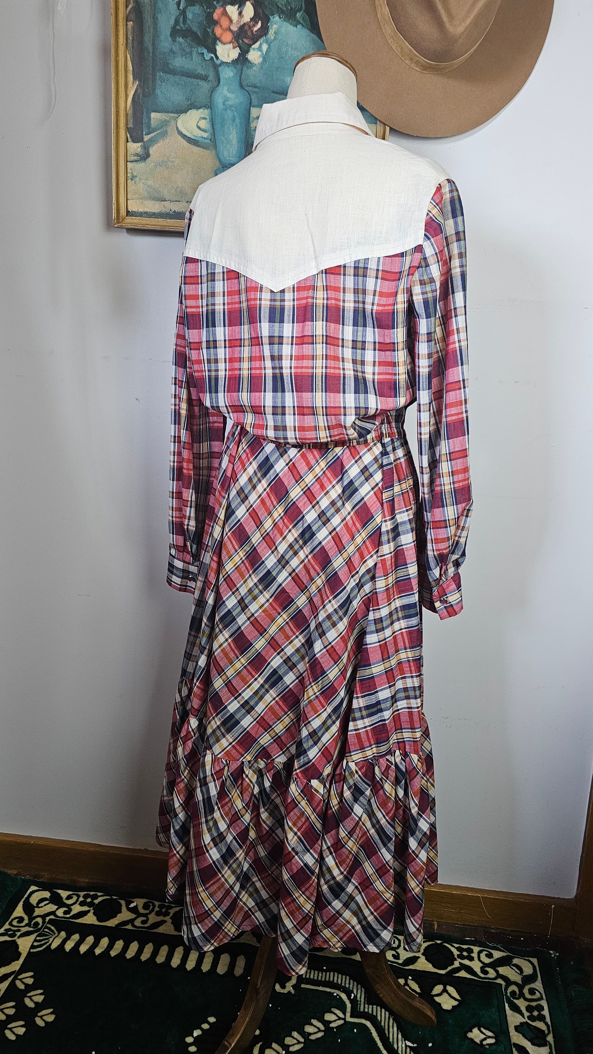 Plaid dress on a mannequin against a white wall with a framed picture and hat in the background.