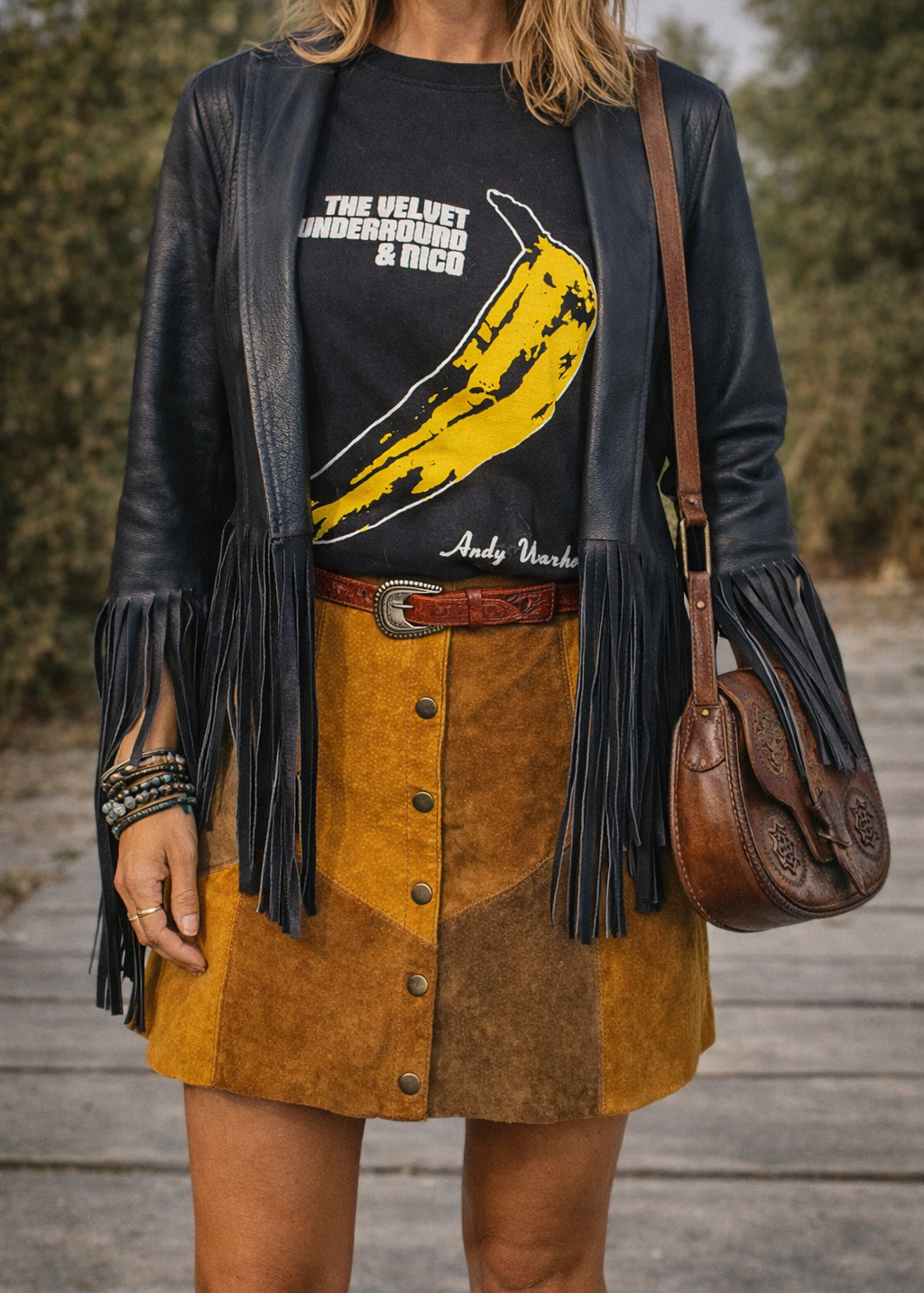 Person wearing a black leather jacket with fringe, a graphic t-shirt, and a brown suede skirt.