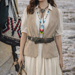 Woman in a beige vest and white dress with a colorful necklace, standing outdoors.