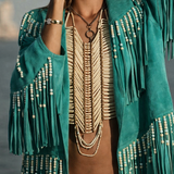 Woman wearing a teal fringe jacket with beads, beige top, and hat against a gray background