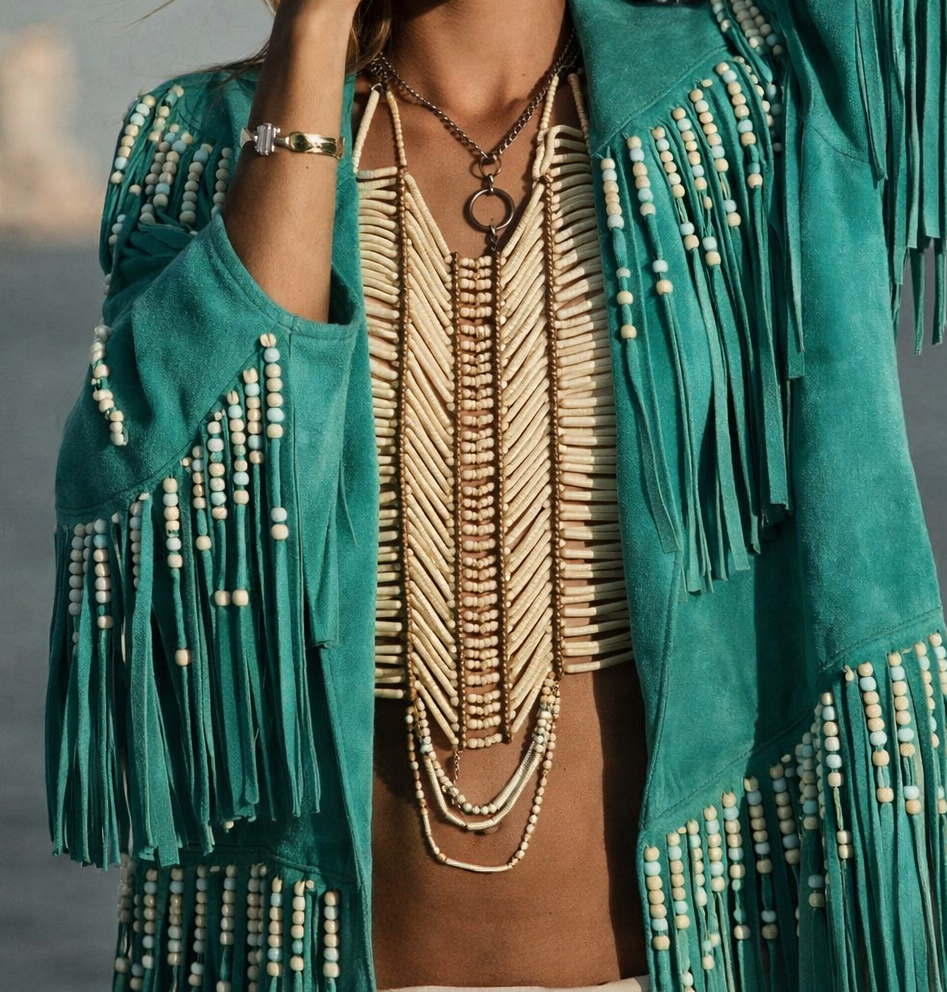 Woman wearing a teal fringe jacket with beads, beige top, and hat against a gray background