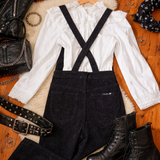 White blouse with black suspenders, black pants, and black boots on a wooden surface.