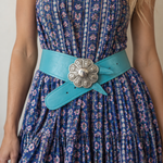A close-up of a woman wearing a blue floral print dress with a wide turquoise belt and a large silver flower-shaped buckle.