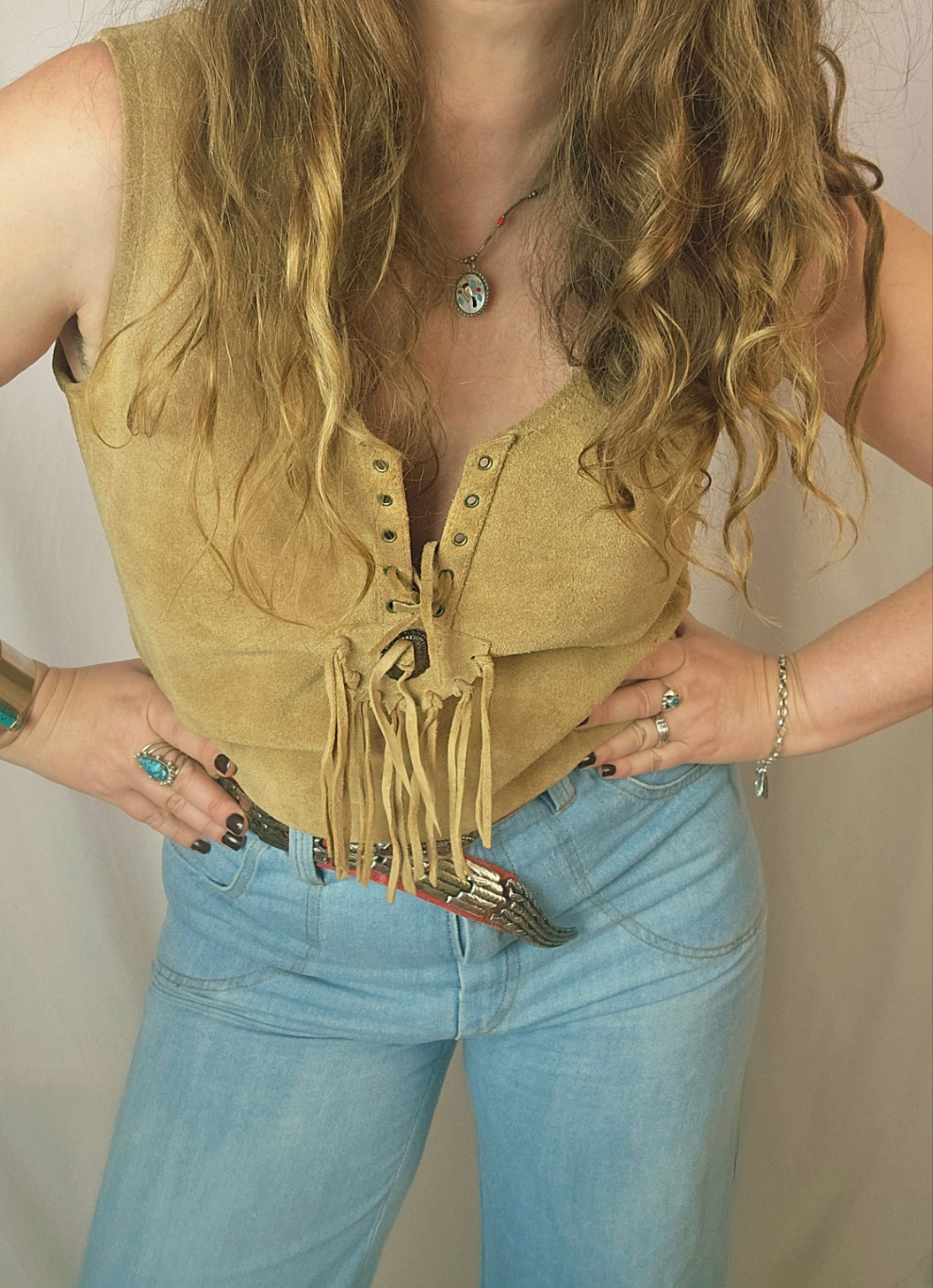 Person wearing a mustard yellow suede vest with fringes and blue jeans against a neutral background