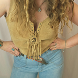 Person wearing a mustard yellow suede vest with fringes and blue jeans against a neutral background