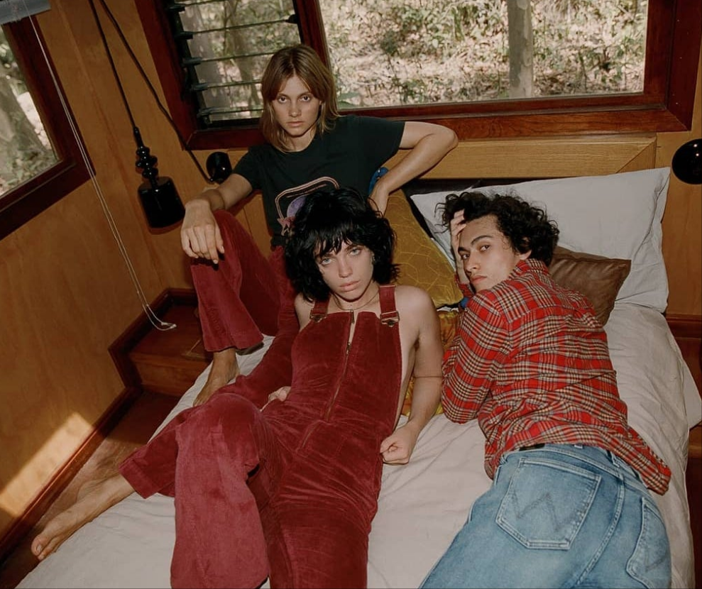 Three people sitting on a bed in a room with wooden walls and a window.