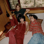 Three people sitting on a bed in a room with wooden walls and a window.