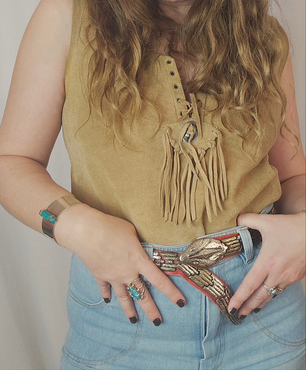 Person wearing a mustard yellow sleeveless top with fringe details and blue jeans, holding a decorative belt.