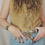 Person wearing a mustard yellow sleeveless top with fringe details and blue jeans, holding a decorative belt.
