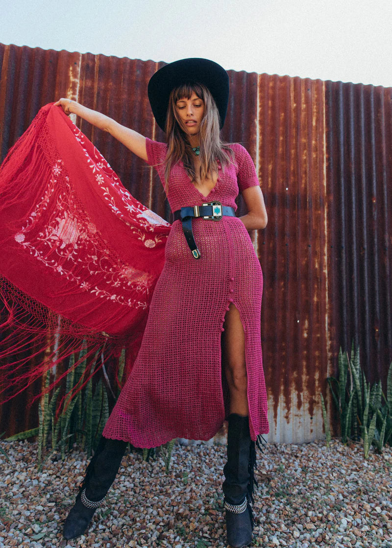 Woman in a pink dress and black hat holding a red scarf against a rustic corrugated metal wall.