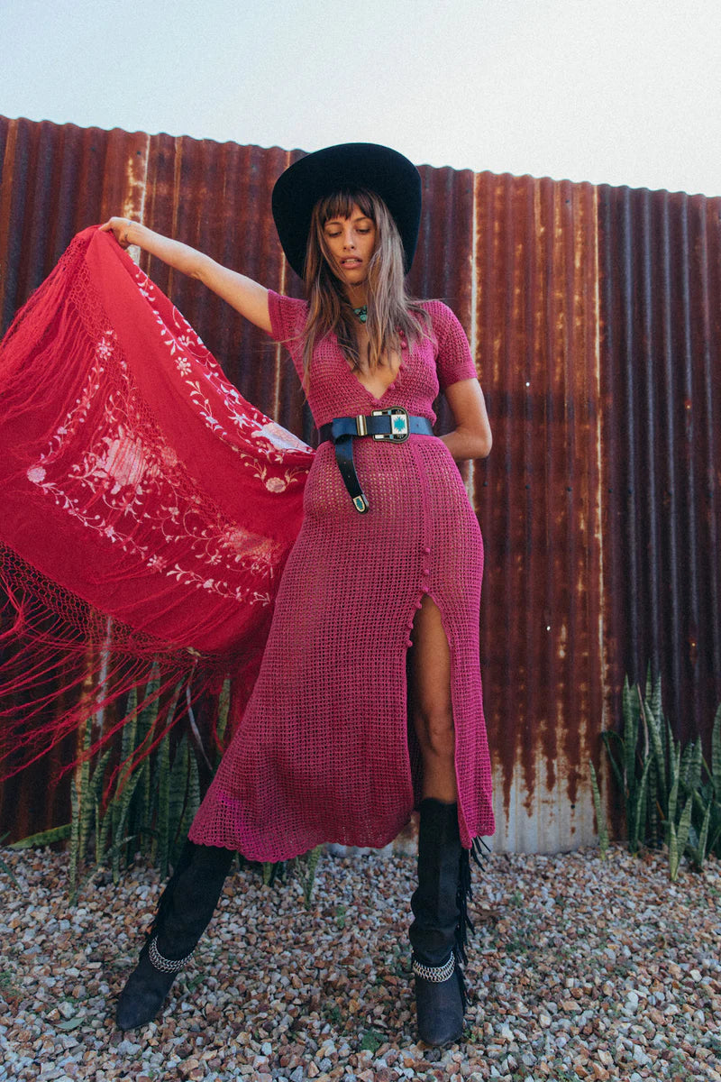Woman in a pink dress and black hat holding a red scarf against a rustic corrugated metal wall.