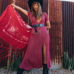 Woman in a pink dress and black hat holding a red scarf against a rustic corrugated metal wall.