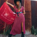 Woman in a pink dress and black hat holding a red scarf against a rustic corrugated metal wall.
