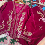 A deep magenta velvet garment, likely a traditional shawl or wrap, featuring intricate gold and light pink paisley embroidery with metallic borders, laid out on a patterned rug.