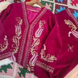 A deep magenta velvet garment, likely a traditional shawl or wrap, featuring intricate gold and light pink paisley embroidery with metallic borders, laid out on a patterned rug.