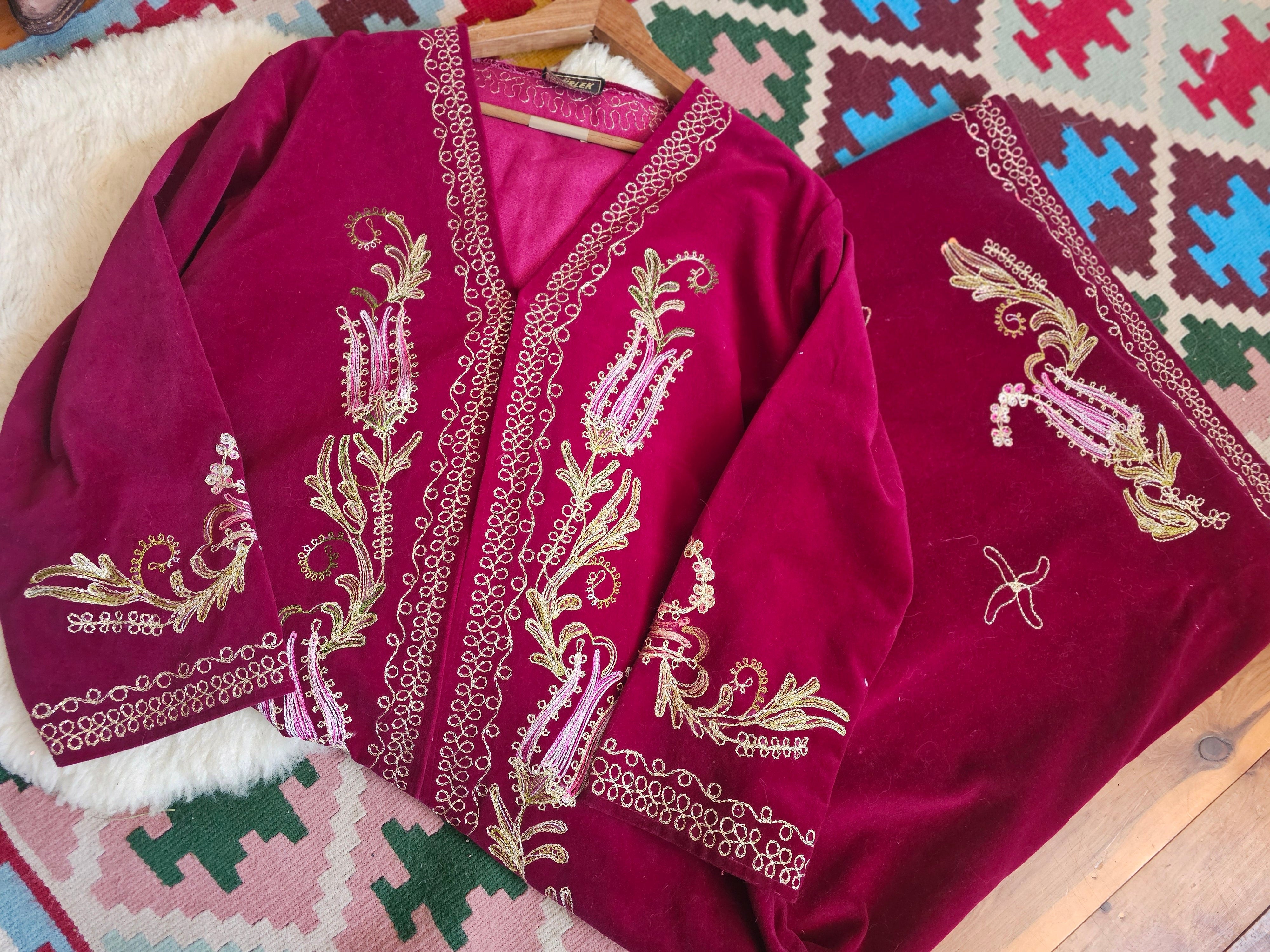 A deep magenta velvet garment, likely a traditional shawl or wrap, featuring intricate gold and light pink paisley embroidery with metallic borders, laid out on a patterned rug.