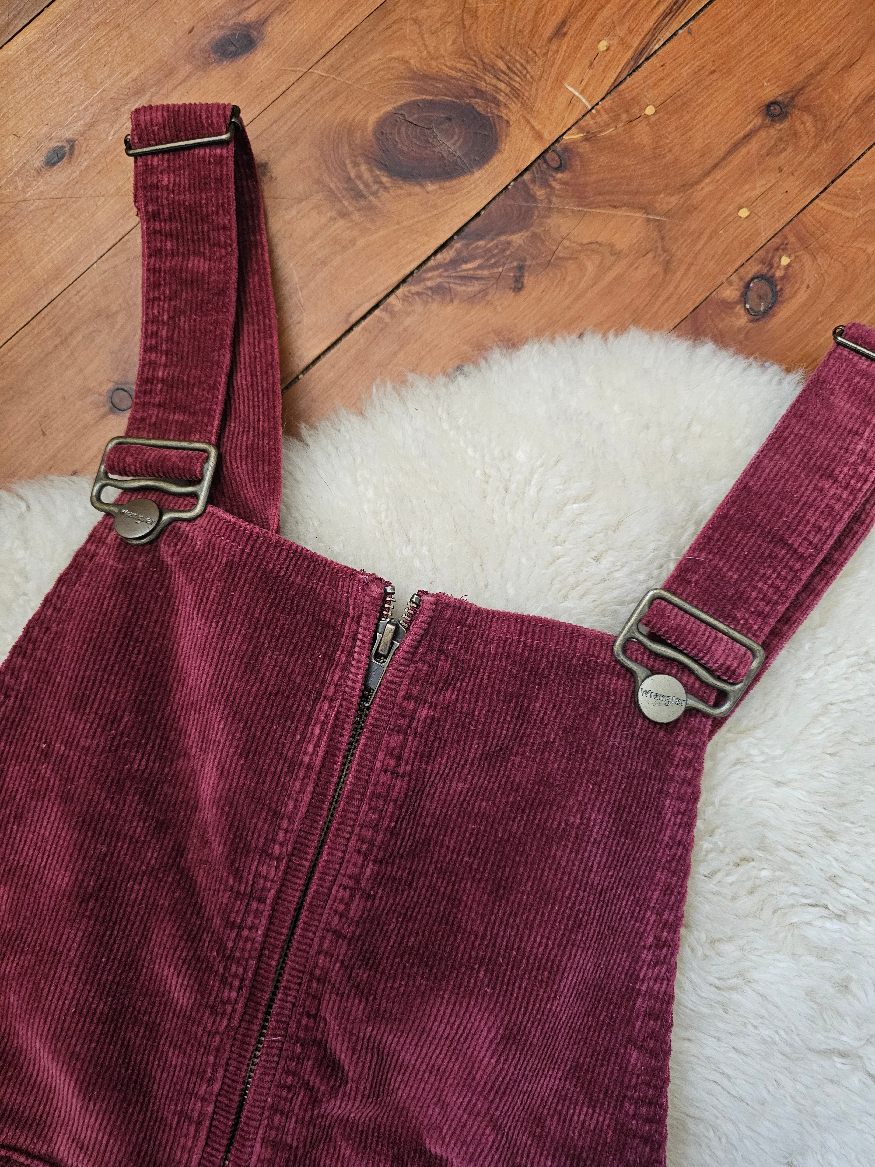 Maroon corduroy overalls with a strap on a wooden floor.
