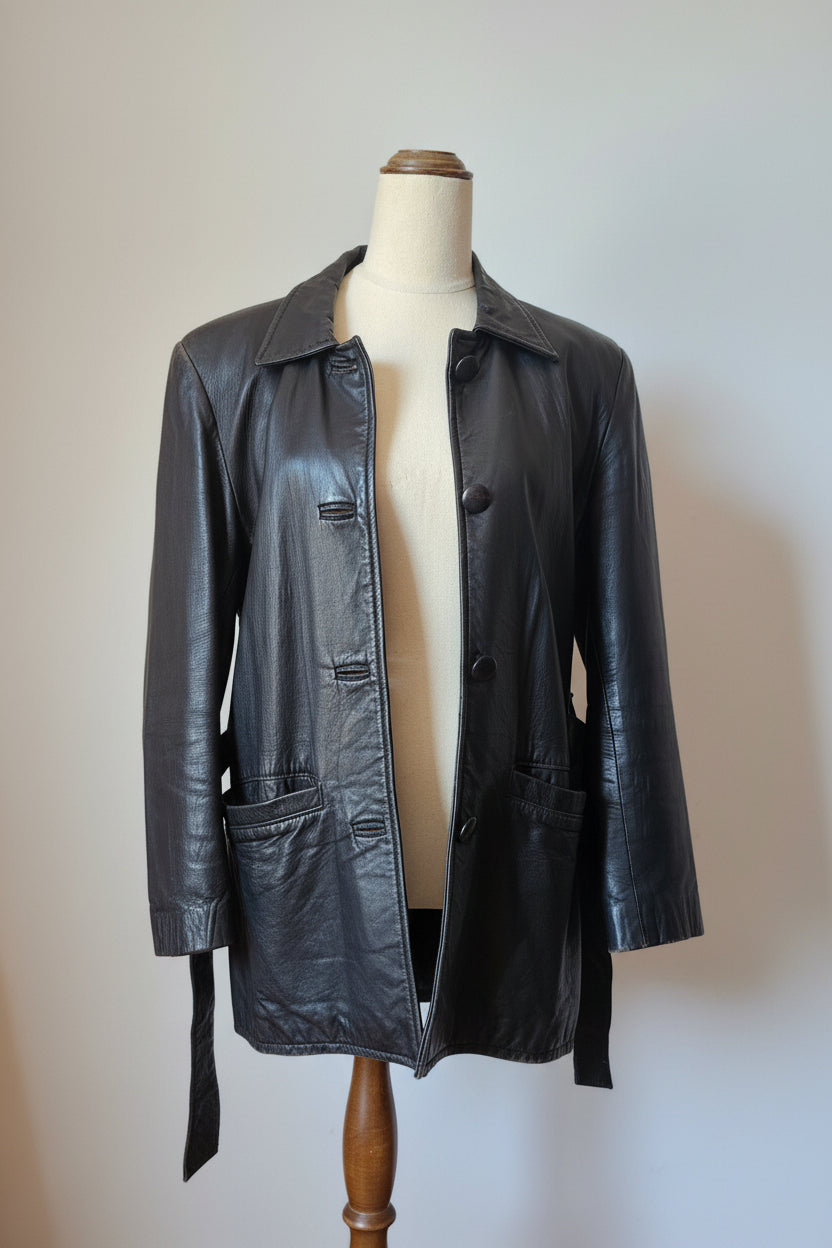 A black leather jacket displayed on a cream-colored mannequin torso, set against a warm beige wall with a wooden wall-hanging decoration in the background.