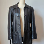 A black leather jacket displayed on a cream-colored mannequin torso, set against a warm beige wall with a wooden wall-hanging decoration in the background.