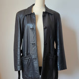 A black leather jacket displayed on a cream-colored mannequin torso, set against a warm beige wall with a wooden wall-hanging decoration in the background.