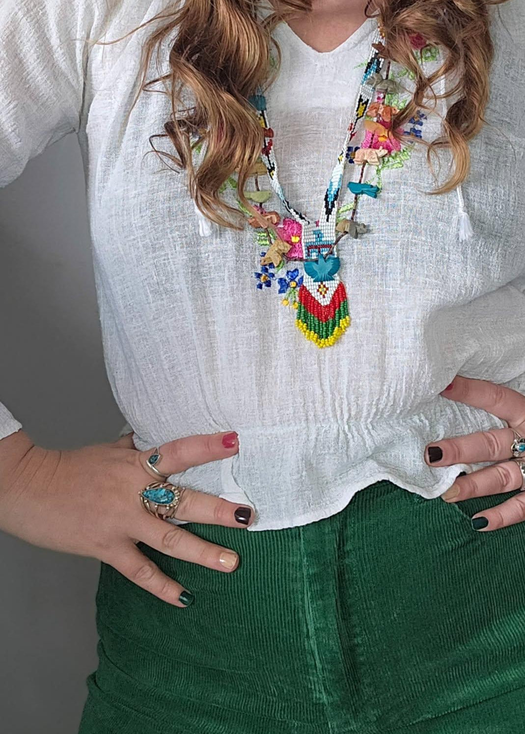 Person wearing a colorful necklace, white blouse, and green pants with a gray background.