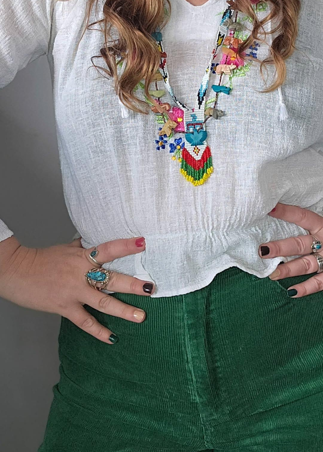 Person wearing a colorful necklace, white blouse, and green pants with a gray background.