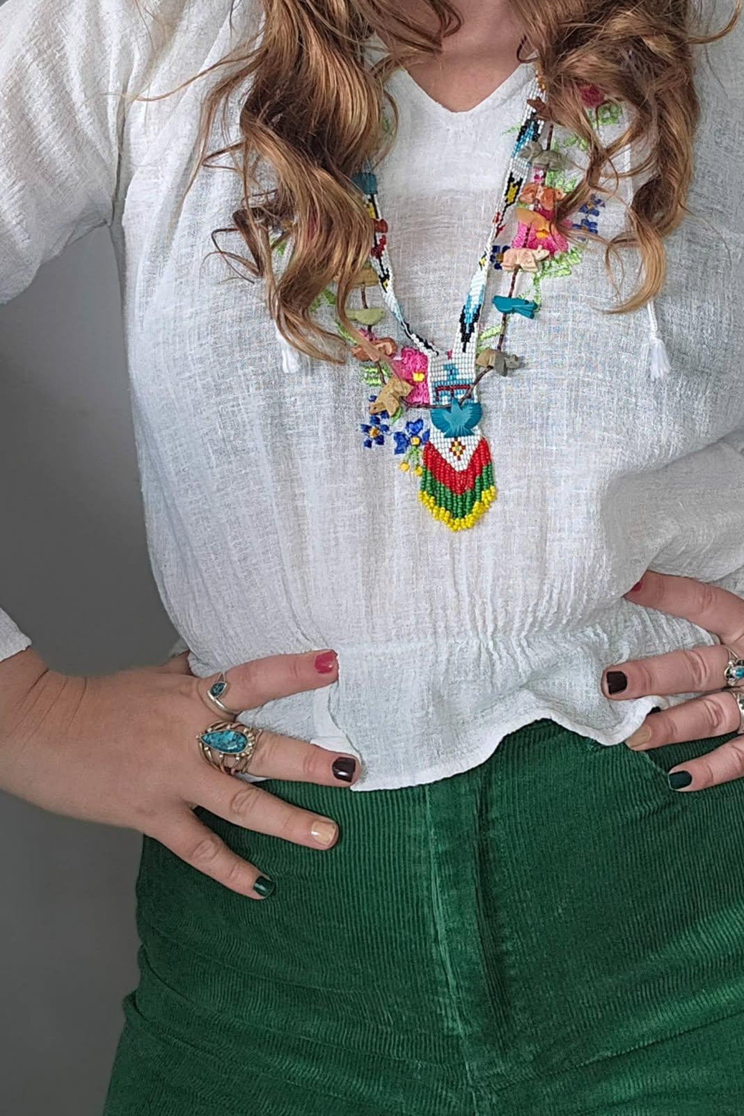 Person wearing a colorful necklace, white blouse, and green pants with a gray background.