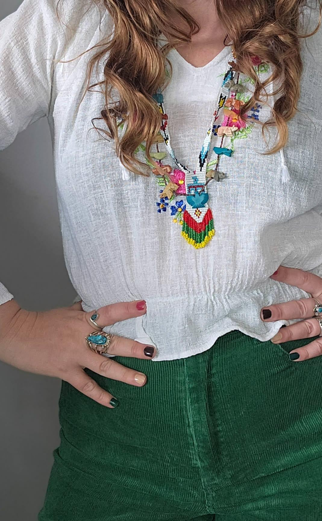 Person wearing a colorful necklace, white blouse, and green pants with a gray background.