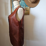 Brown leather bag with a beige interior on a white surface, with a hat and framed picture in the background.