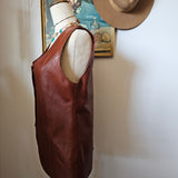 Brown leather bag with a beige interior on a white surface, with a hat and framed picture in the background.