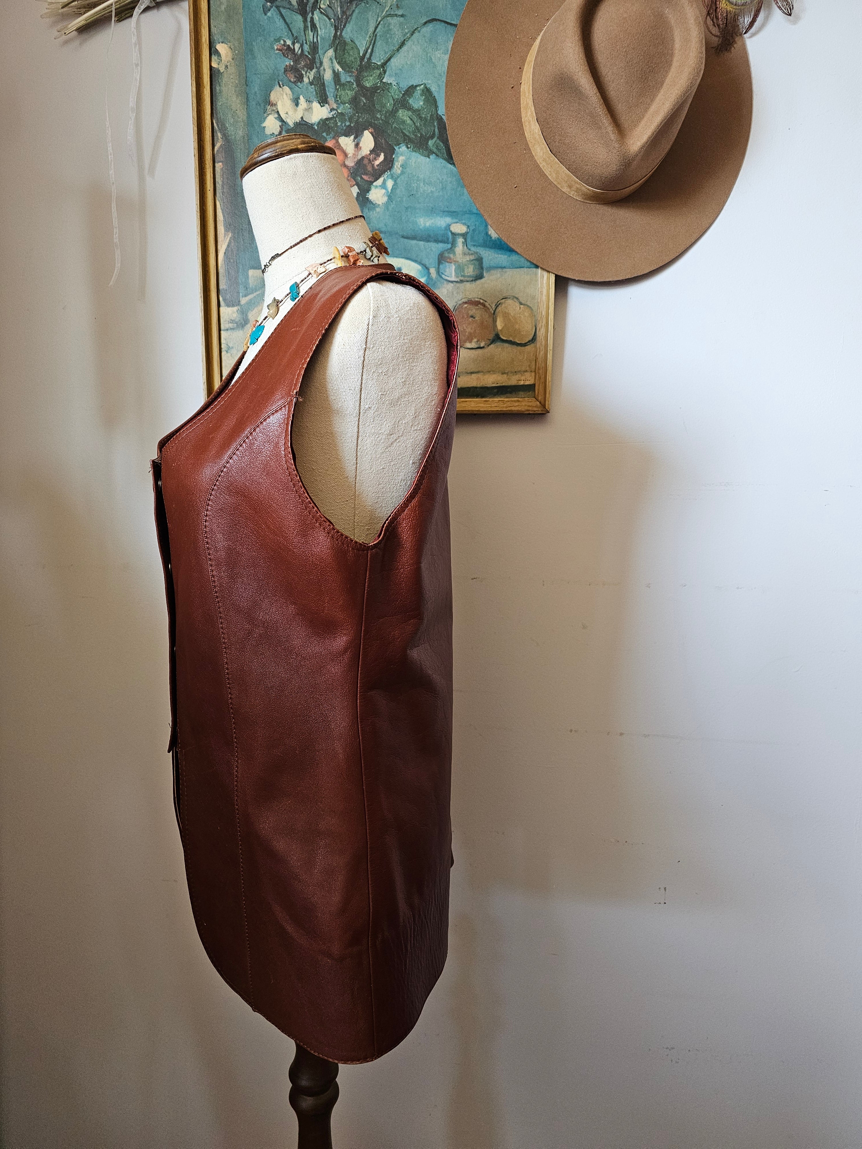Brown leather bag with a beige interior on a white surface, with a hat and framed picture in the background.