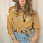 Woman wearing a beige embroidered jacket, blue jeans, and a brown hat on a plain background