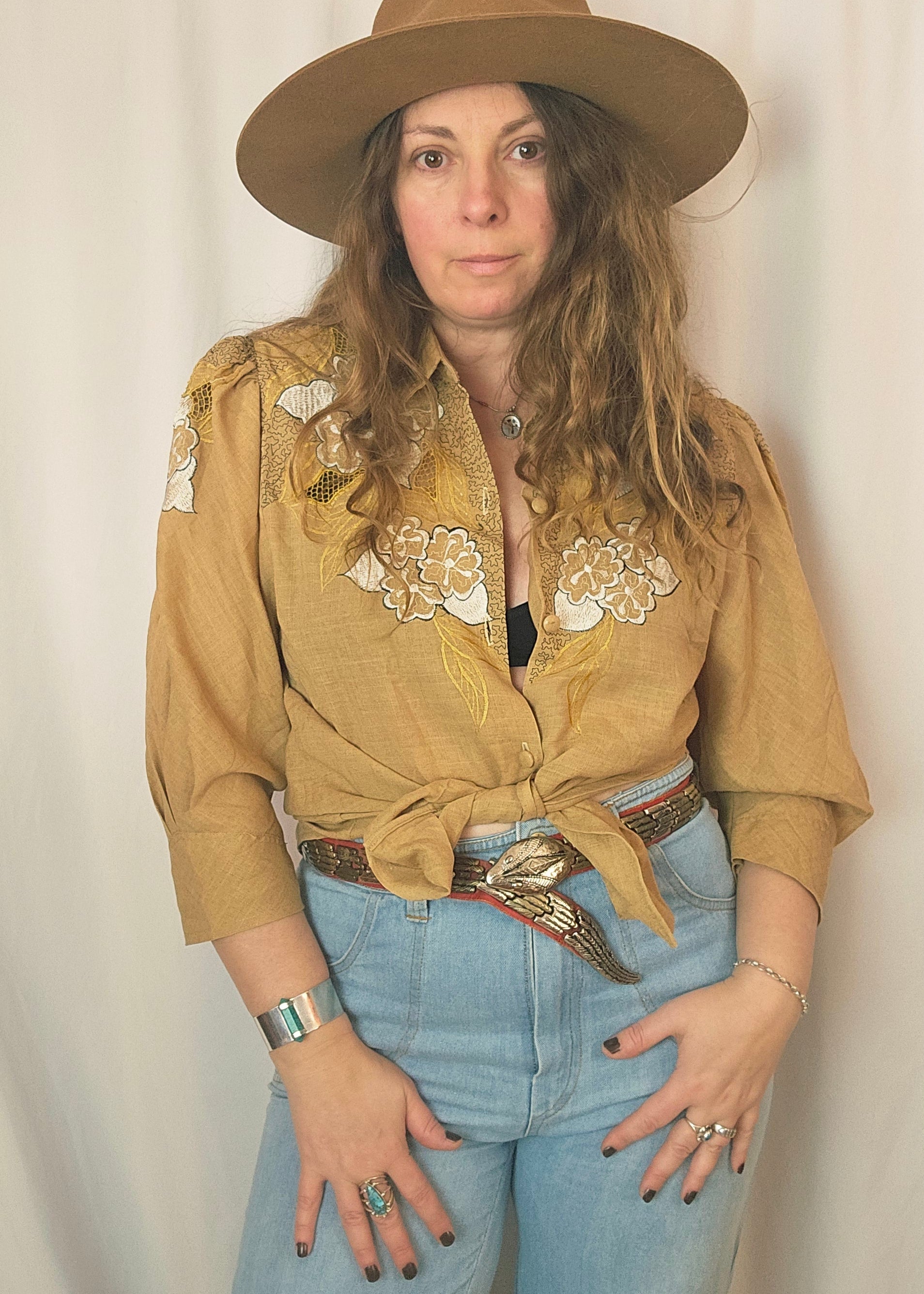 Woman wearing a beige embroidered jacket, blue jeans, and a brown hat on a plain background