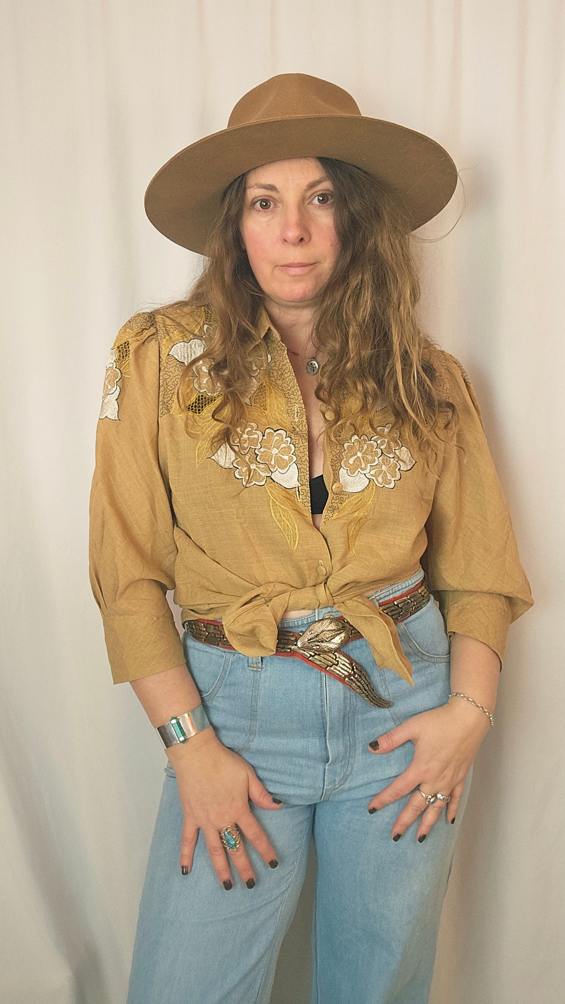 Woman wearing a beige embroidered jacket, blue jeans, and a brown hat on a plain background