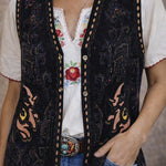 Person wearing a black embroidered vest over a white blouse with floral design, paired with blue jeans.